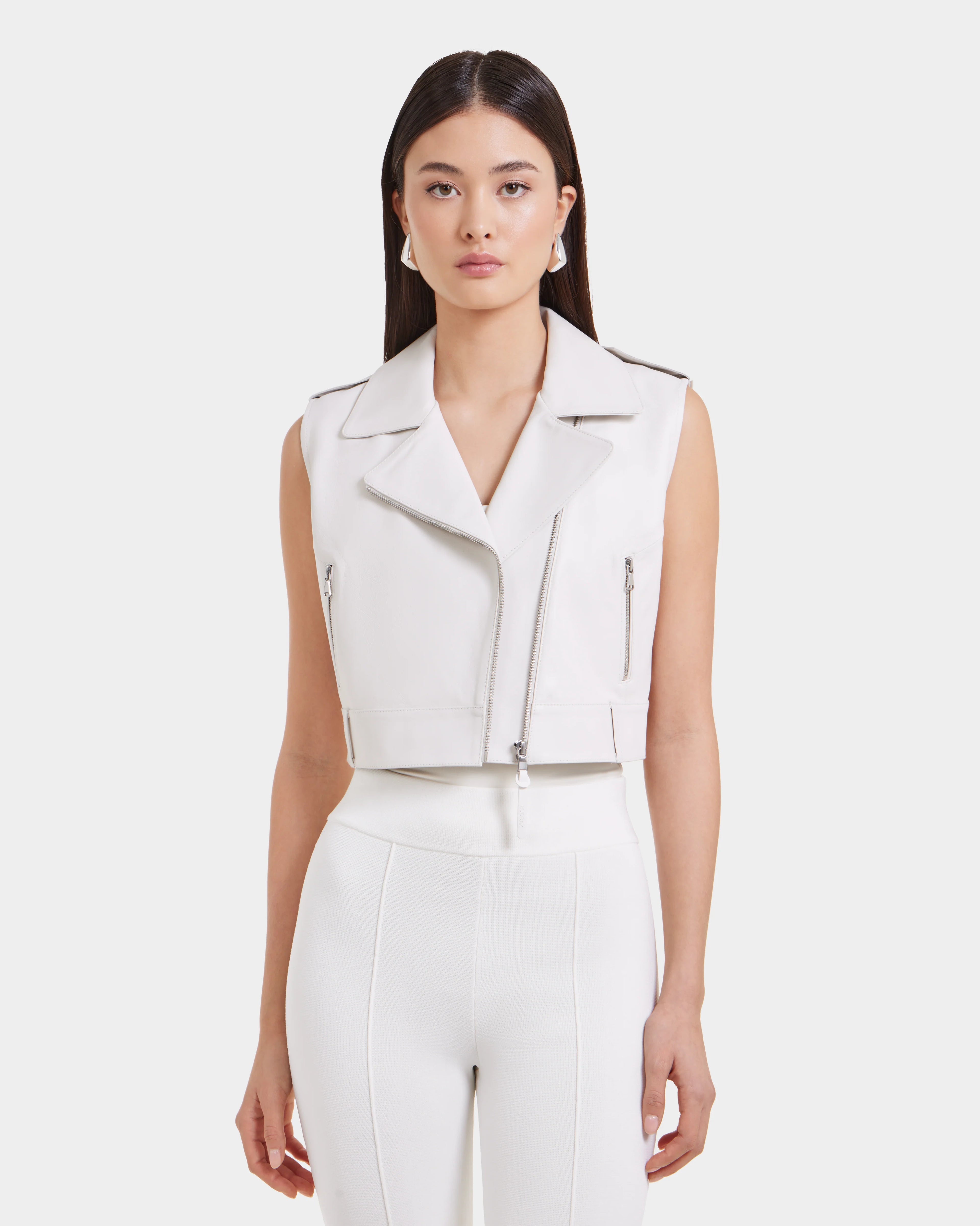 A woman with straight dark hair models the Rudsak BETTY - 8125000 Optic White high-waisted pants and a cropped moto leather vest, standing against a light background for a modern, cool vibe.