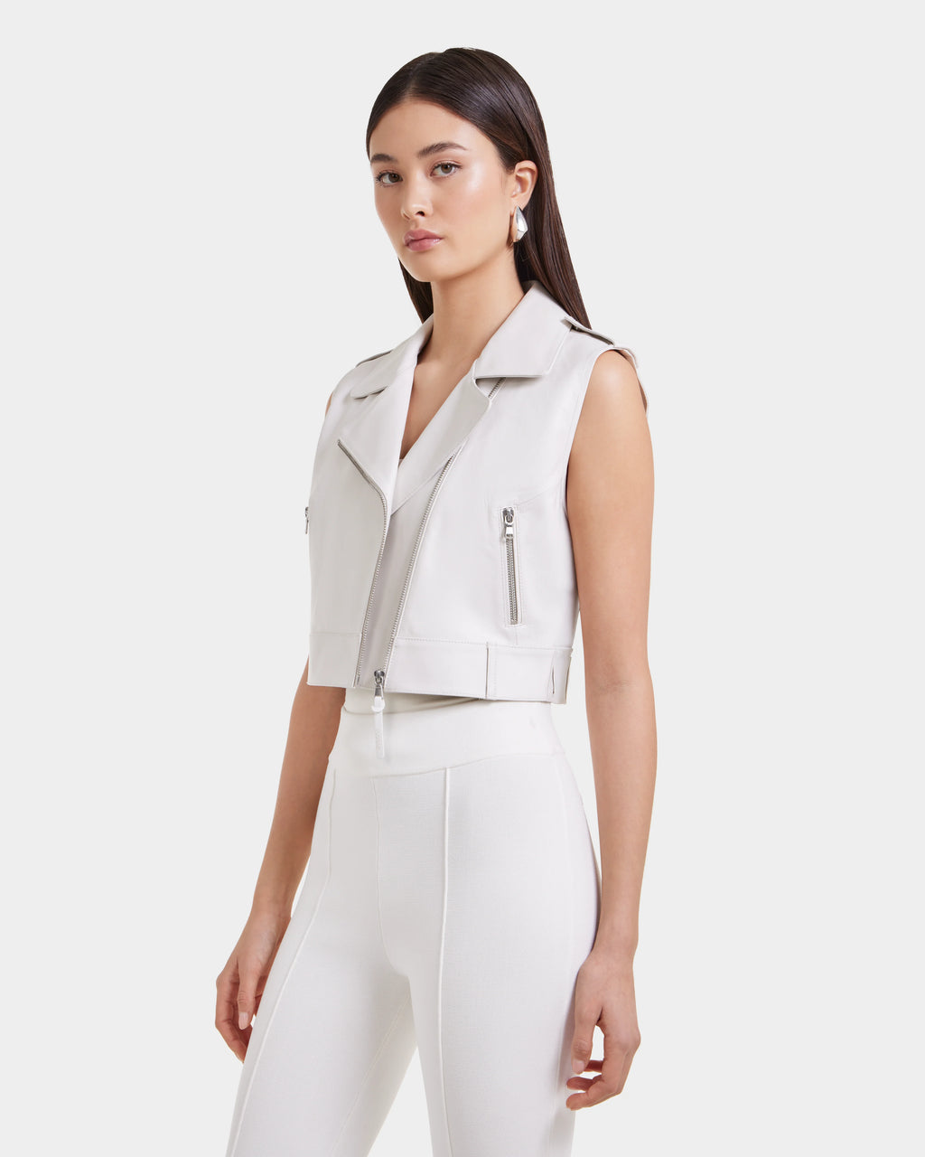 A woman with straight dark hair models the Rudsak BETTY - 8125000 Optic White sleeveless moto vest with zippered pockets and high-waisted white pants, standing against a plain light backdrop.
