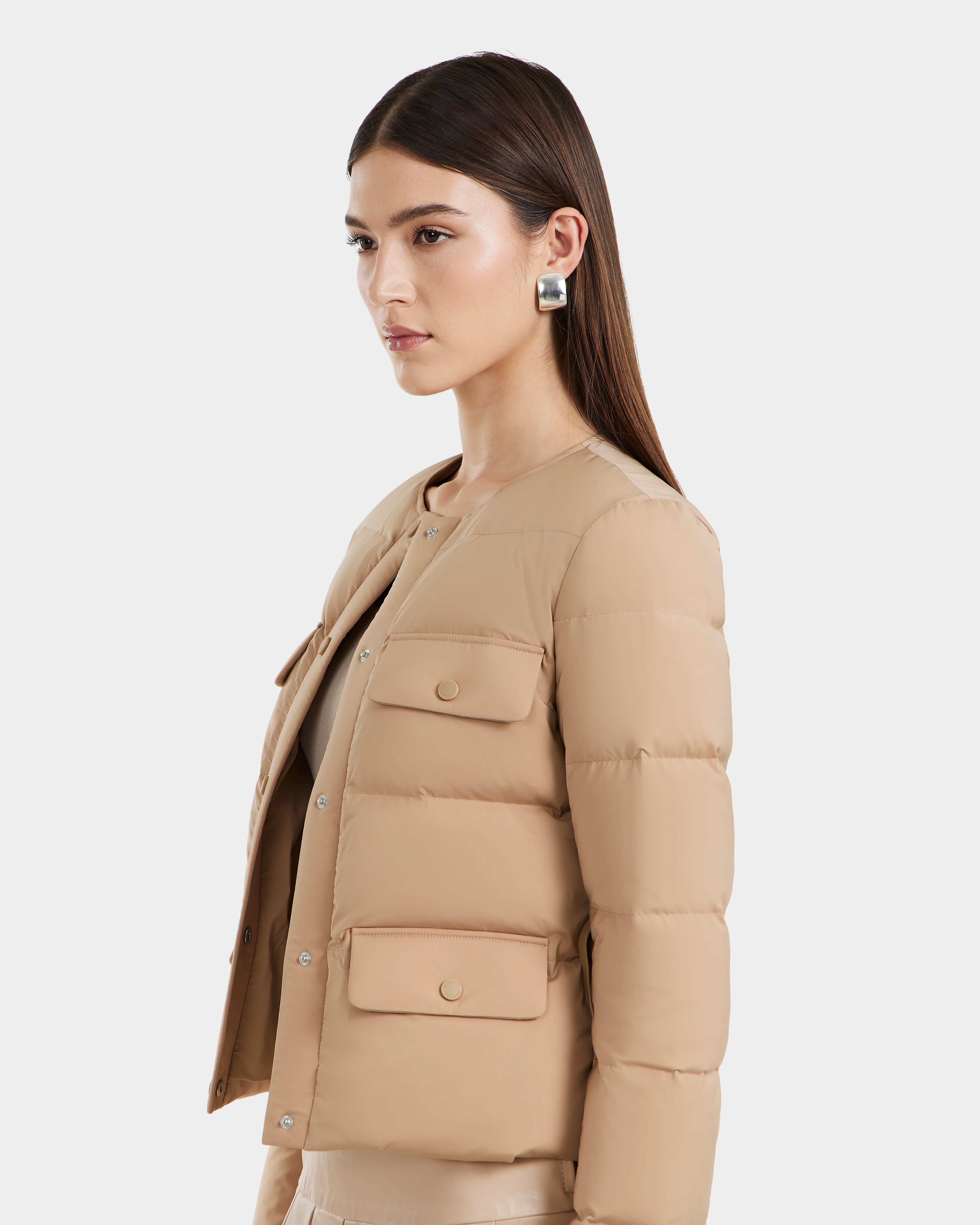 A woman with straight brown hair models the Rudsak FRANCESCA - 8125021 Almond quilted puffer jacket, featuring flap pockets and silver statement earrings, posed against a white background.