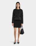 A woman with straight dark hair and a neutral expression stands against a plain background wearing the Rudsak FRANCESCA - 8125021 Black RDS-certified down puffer jacket with a belt, paired with a black skirt, heels, and handbag.