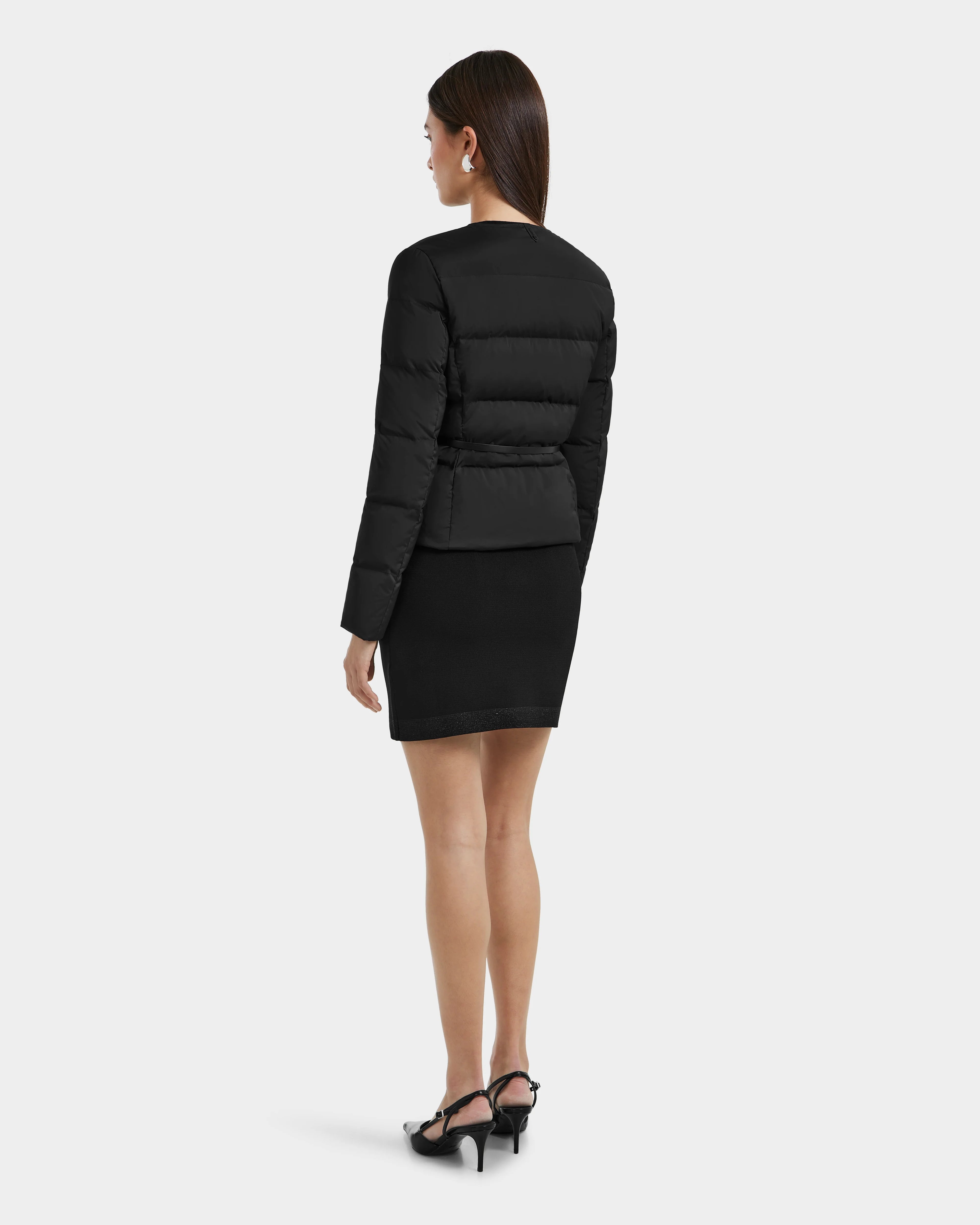 A woman faces away, wearing the Rudsak FRANCESCA - 8125021 Black puffer jacket with a vegan leather belt, paired with a black knee-length skirt and heeled sandals against a plain light background.