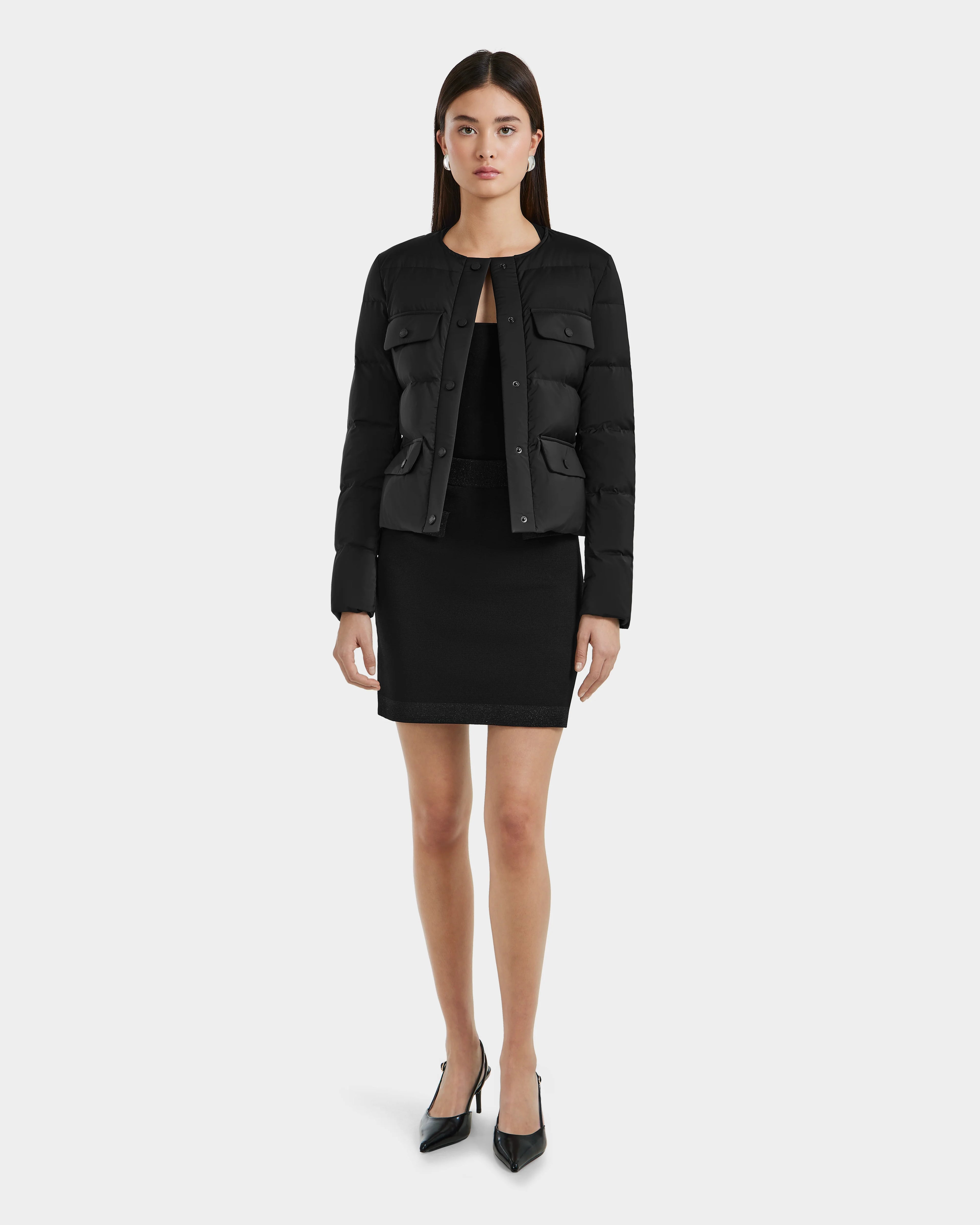 A woman faces forward against a plain white background, wearing the Rudsak FRANCESCA - 8125021 Black cropped jacket, paired with a black top, black skirt, and black pointed heels.