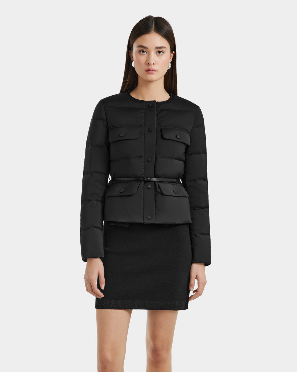 A woman with straight dark hair models the Rudsak FRANCESCA - 8125021 Black lightweight puffer jacket, featuring buttons and front pockets, paired with a matching black skirt against a plain light background.