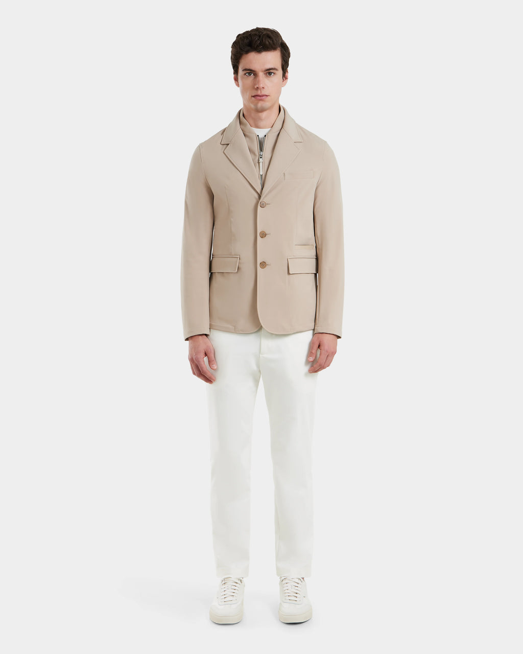 A man stands facing forward in the KEITH - 6125045 Almond blazer-style jacket by Rudsak, worn over a light zip-up top with white trousers and sneakers against a plain background—a modern, refined addition to any mens wardrobe.