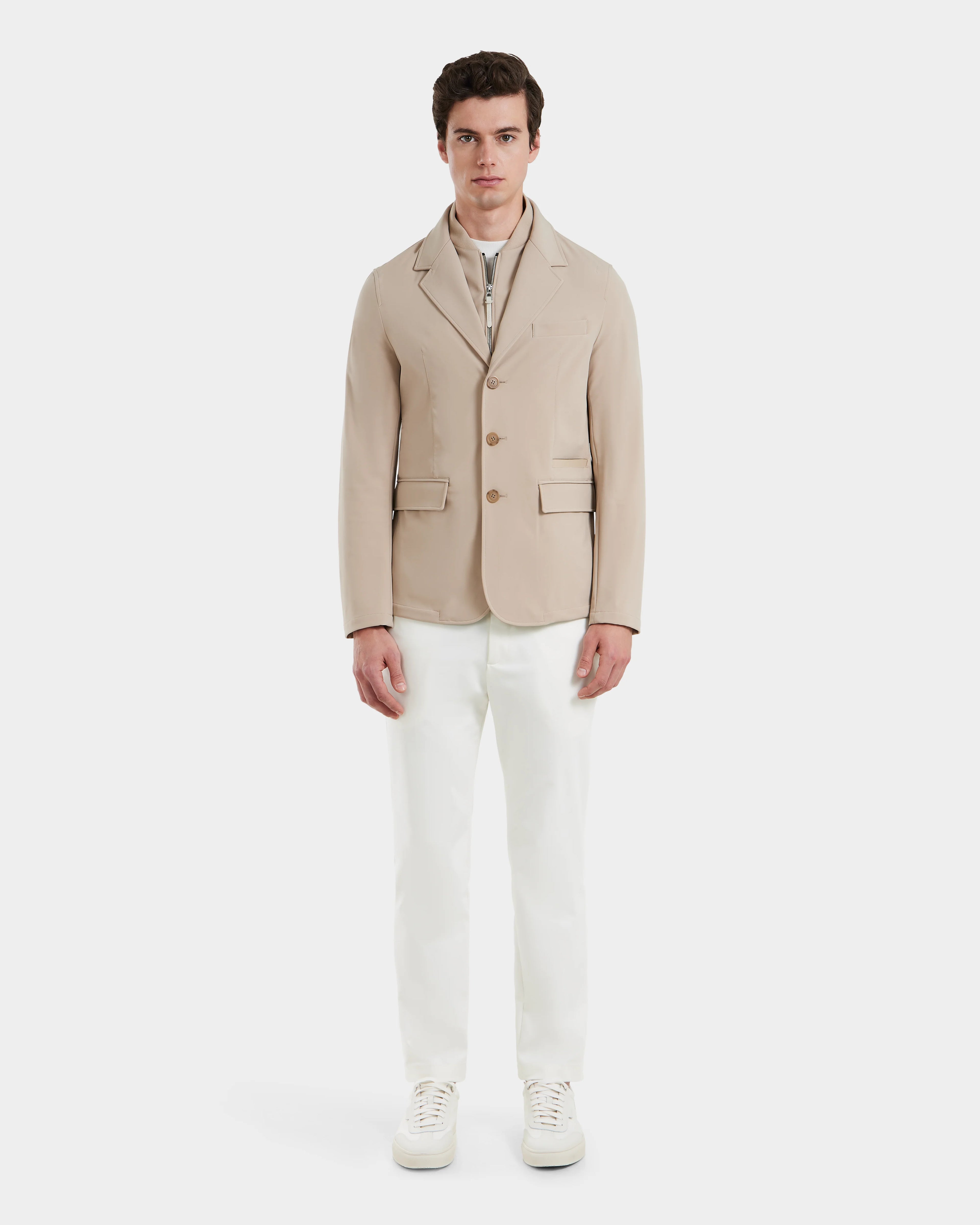 A man stands facing forward in the KEITH - 6125045 Almond blazer-style jacket by Rudsak, worn over a light zip-up top with white trousers and sneakers against a plain background—a modern, refined addition to any mens wardrobe.