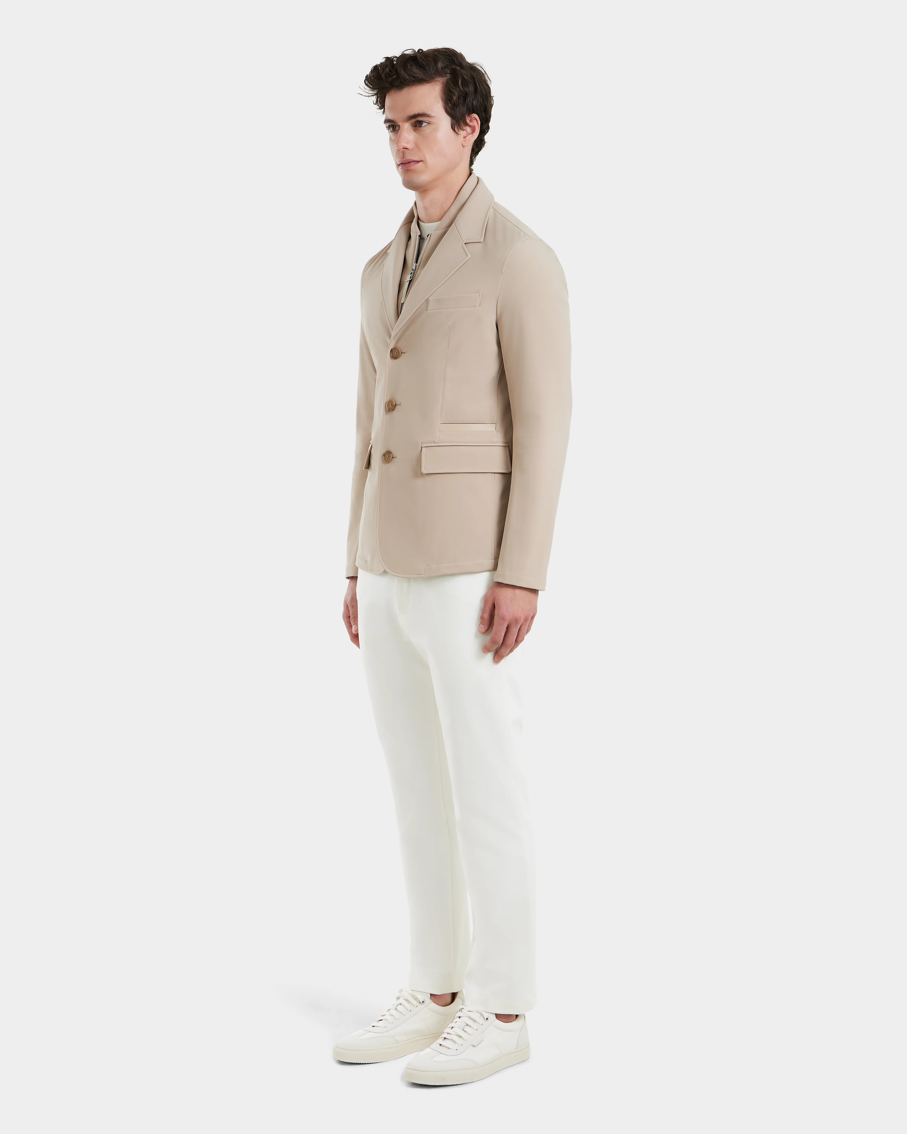 A man faces slightly left, wearing the KEITH - 6125045 Almond blazer-style jacket by Rudsak over a light shirt, white pants, and white sneakers against a plain background—highlighting modern tailoring for men.