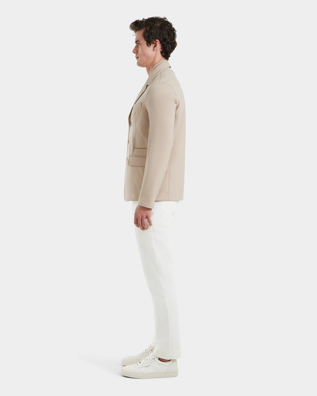 A man stands in profile against a white background, showcasing Rudsaks KEITH - 6125045 Almond blazer-style jacket, paired with white pants and sneakers—an effortless addition to any modern mens wardrobe.