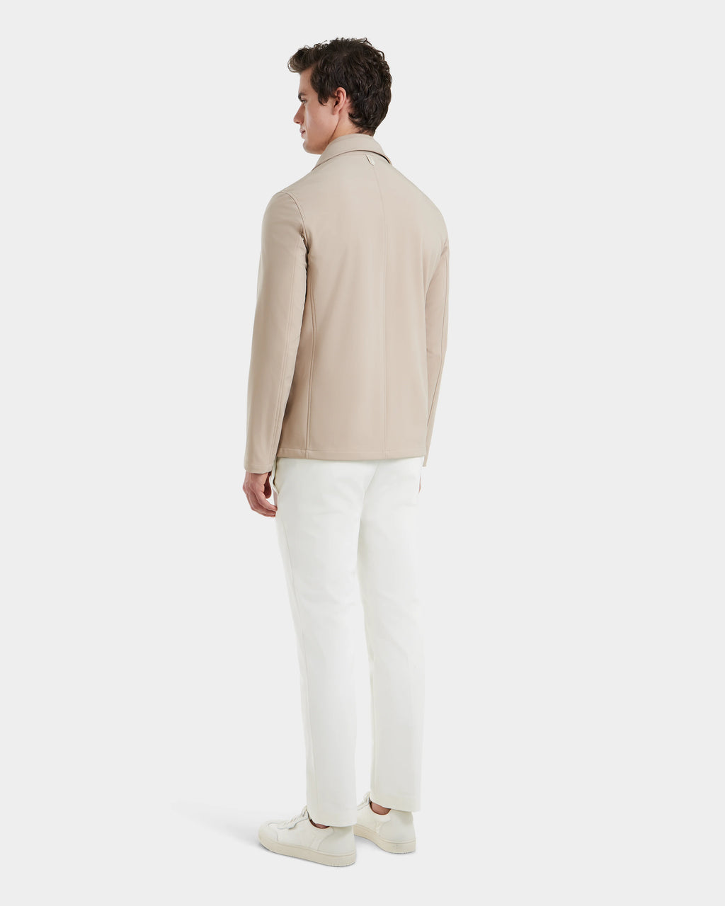 A man faces away, wearing the Rudsak KEITH - 6125045 Almond beige blazer-style jacket with modern tailoring, paired with white pants and white sneakers against a plain white background.