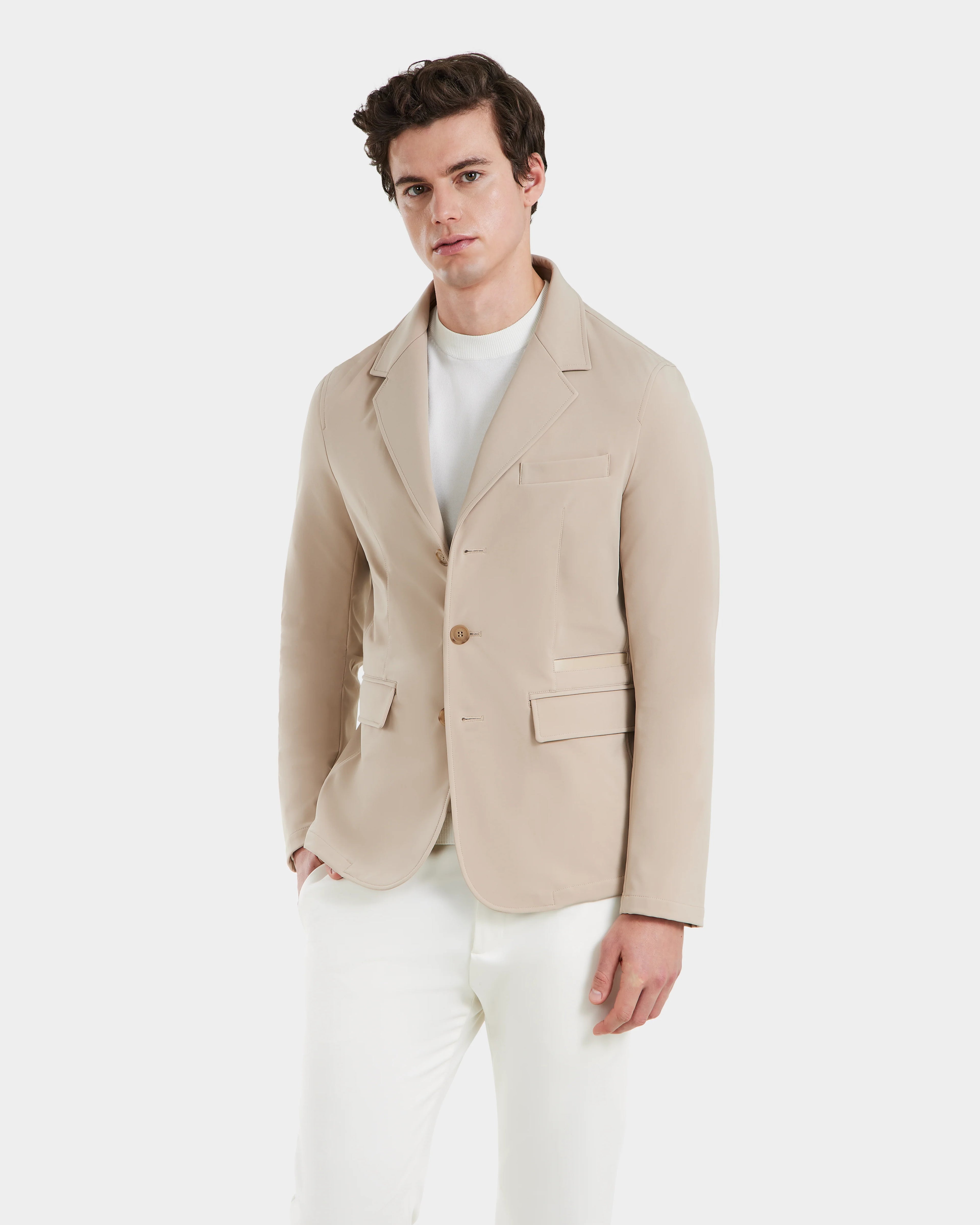 A man models the Rudsak KEITH - 6125045 Almond blazer-style jacket over a white shirt and pants, standing against a plain light background and gazing at the camera with a neutral expression.