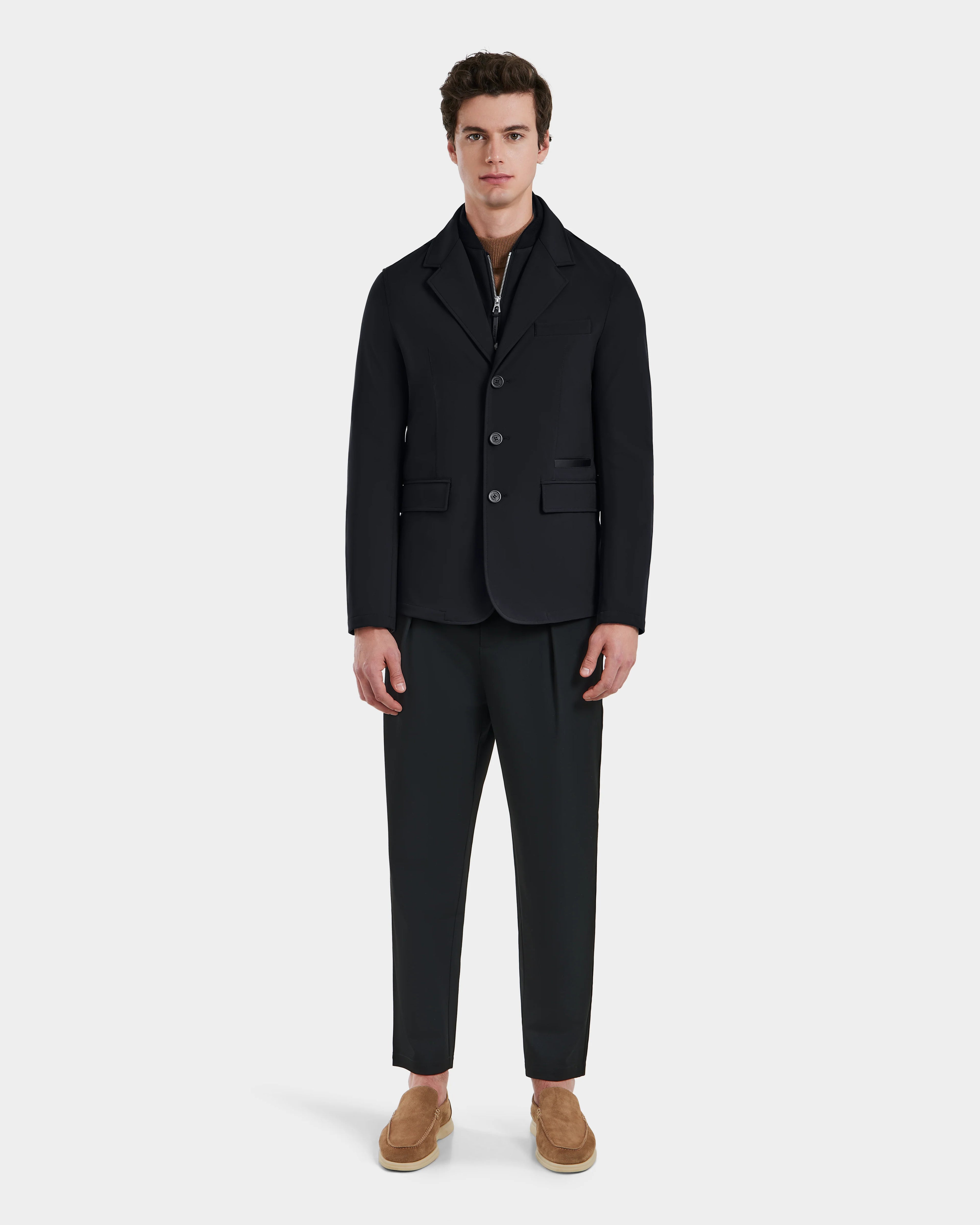 A man stands against a plain background, wearing Rudsak’s KEITH - 6125045 Black jacket with matching trousers, a zip-up shirt, and light brown slip-ons—a refined look that showcases modern tailoring in versatile menswear.