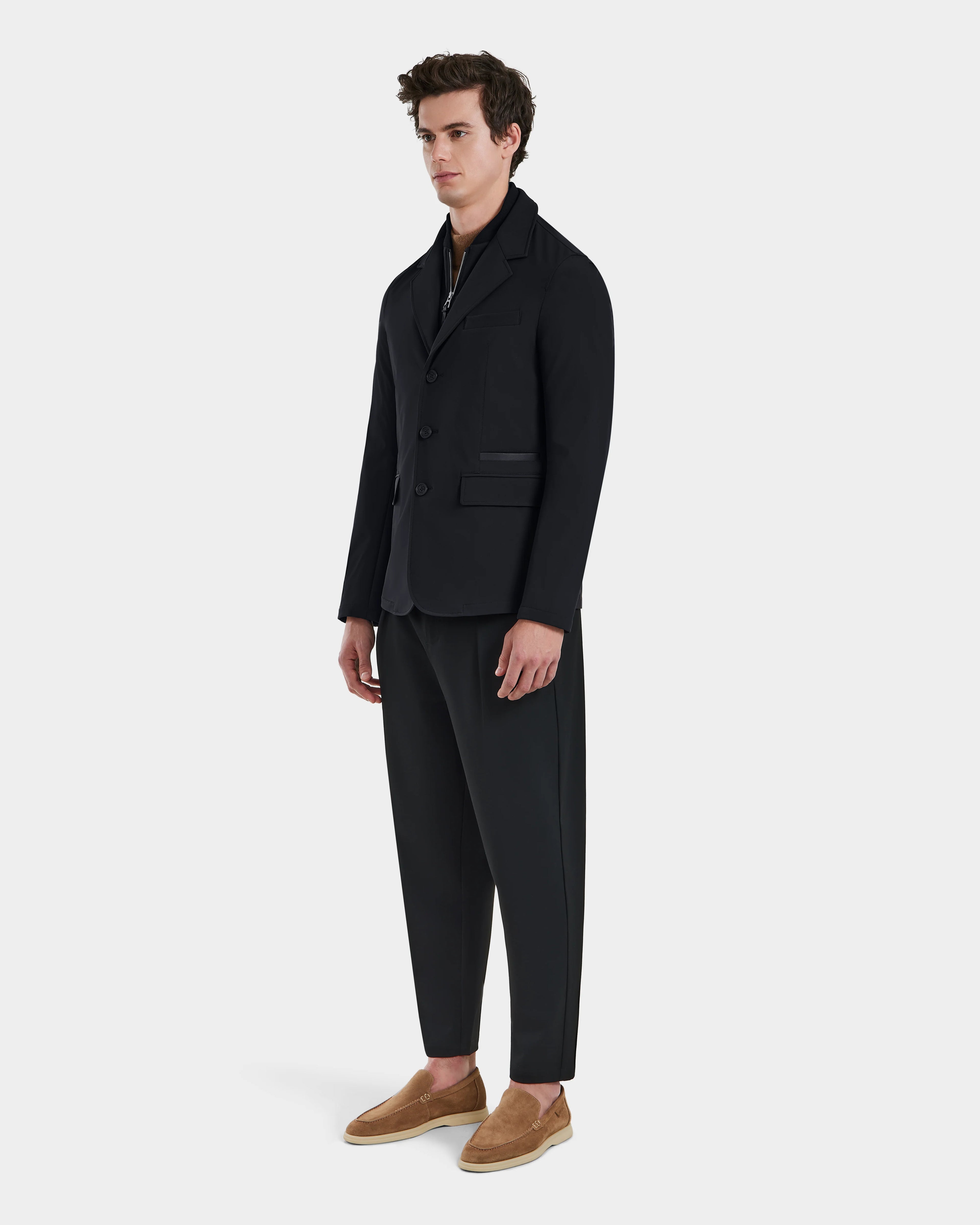 A man stands against a plain light background, wearing the Rudsak KEITH - 6125045 Black blazer-style jacket and matching trousers, paired with a dark shirt and tan slip-on shoes. He looks forward neutrally, showcasing modern tailoring.
