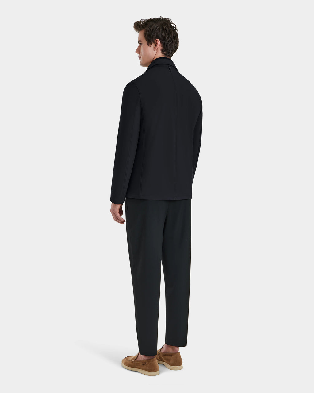 A man faces away wearing the Rudsak KEITH - 6125045 Black blazer-style jacket, paired with black pants and tan slip-on shoes, set against a plain light gray background—showcasing modern, versatile menswear.