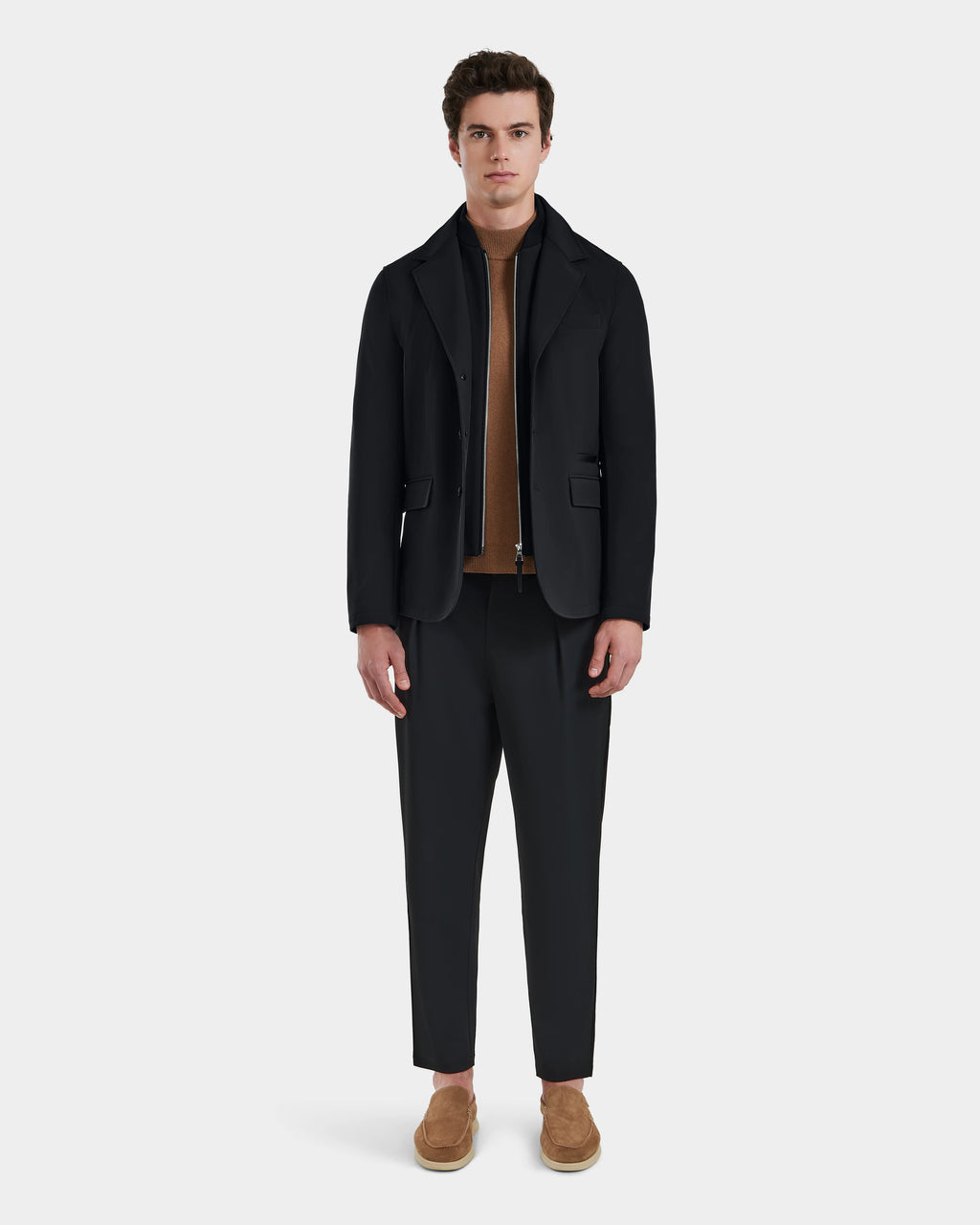 A man faces forward in the refined KEITH - 6125045 Black blazer-style jacket by Rudsak, worn over a brown sweater with black trousers and light brown slip-on shoes. A plain, light background highlights this versatile menswear look.