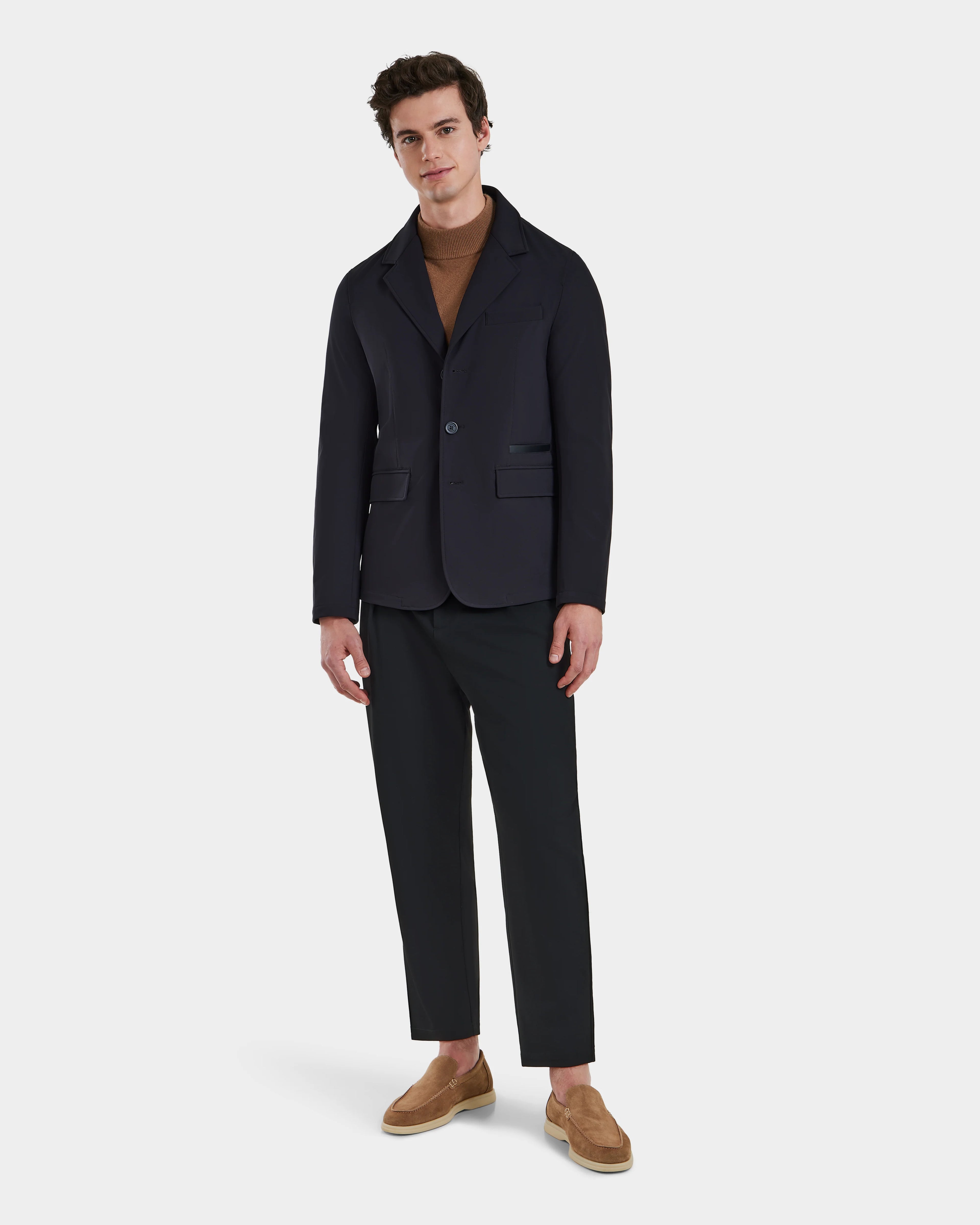 A man with short dark hair stands against a white background wearing the KEITH - 6125045 Black blazer-style jacket by Rudsak, paired with a brown turtleneck, black pants, and tan slip-on shoes for a modern, versatile menswear look.