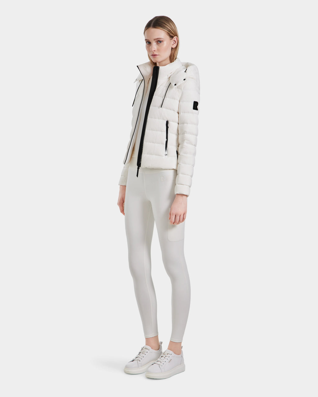 A woman stands against a plain white background, wearing the Rudsak MELISSA - 8124031 Cotton White lightweight down puffer, white turtleneck, fitted white leggings, and white sneakers. She faces forward with a neutral expression.