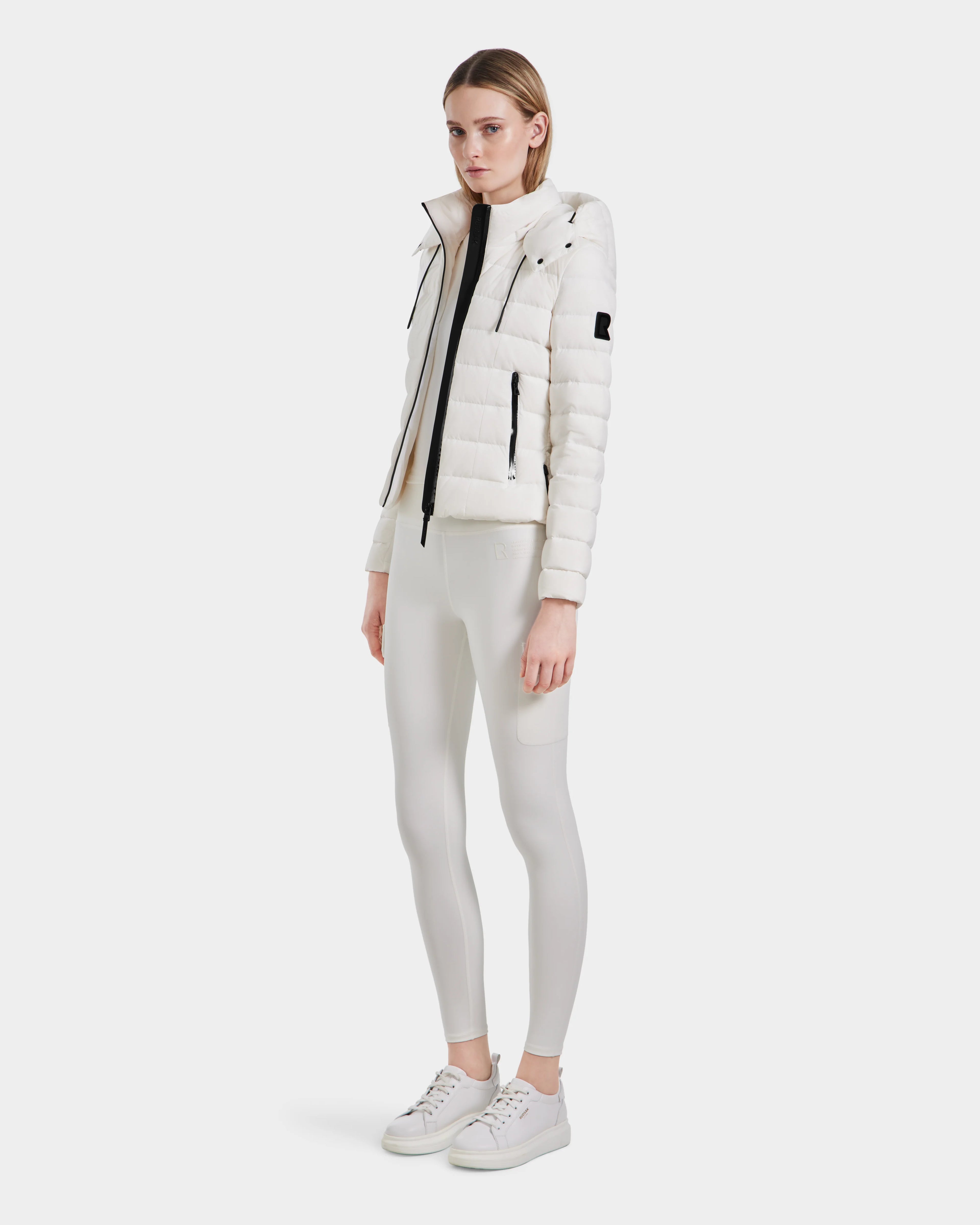 A woman stands against a plain white background, wearing the Rudsak MELISSA - 8124031 Cotton White lightweight down puffer, white turtleneck, fitted white leggings, and white sneakers. She faces forward with a neutral expression.