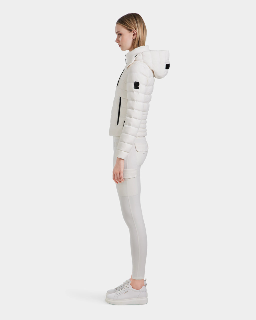 A woman in profile wears the Rudsak MELISSA - 8124031 Cotton White lightweight down puffer with RDS certified down, paired with matching pants and white sneakers, set against a plain white background.