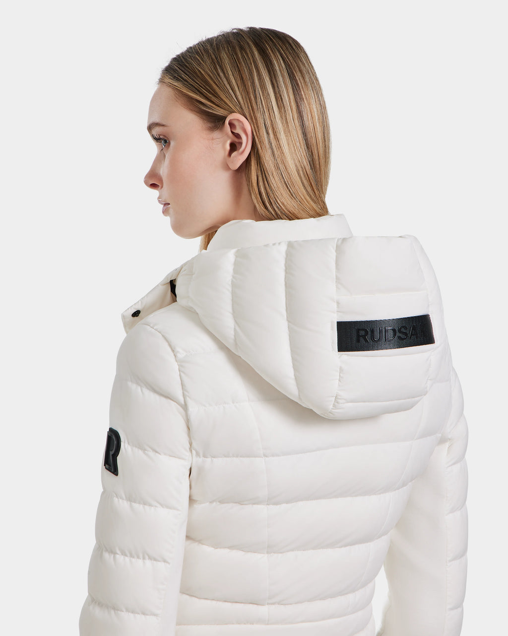 A woman with straight blonde hair wears the Rudsak MELISSA - 8124031 white quilted womens lightweight down puffer jacket with a hood, featuring a black RUDSAK label on the back. She is facing away, looking left.