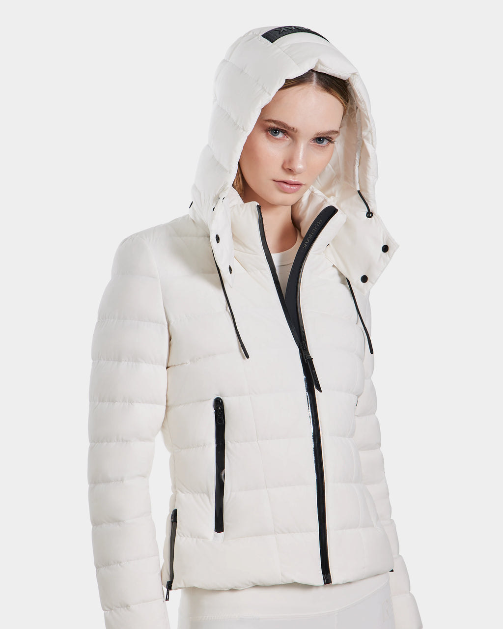 A woman wears the Rudsak MELISSA - 8124031 Cotton White quilted hooded lightweight down puffer with black zippers, standing confidently against a plain white background, her hood up for added coziness and style.