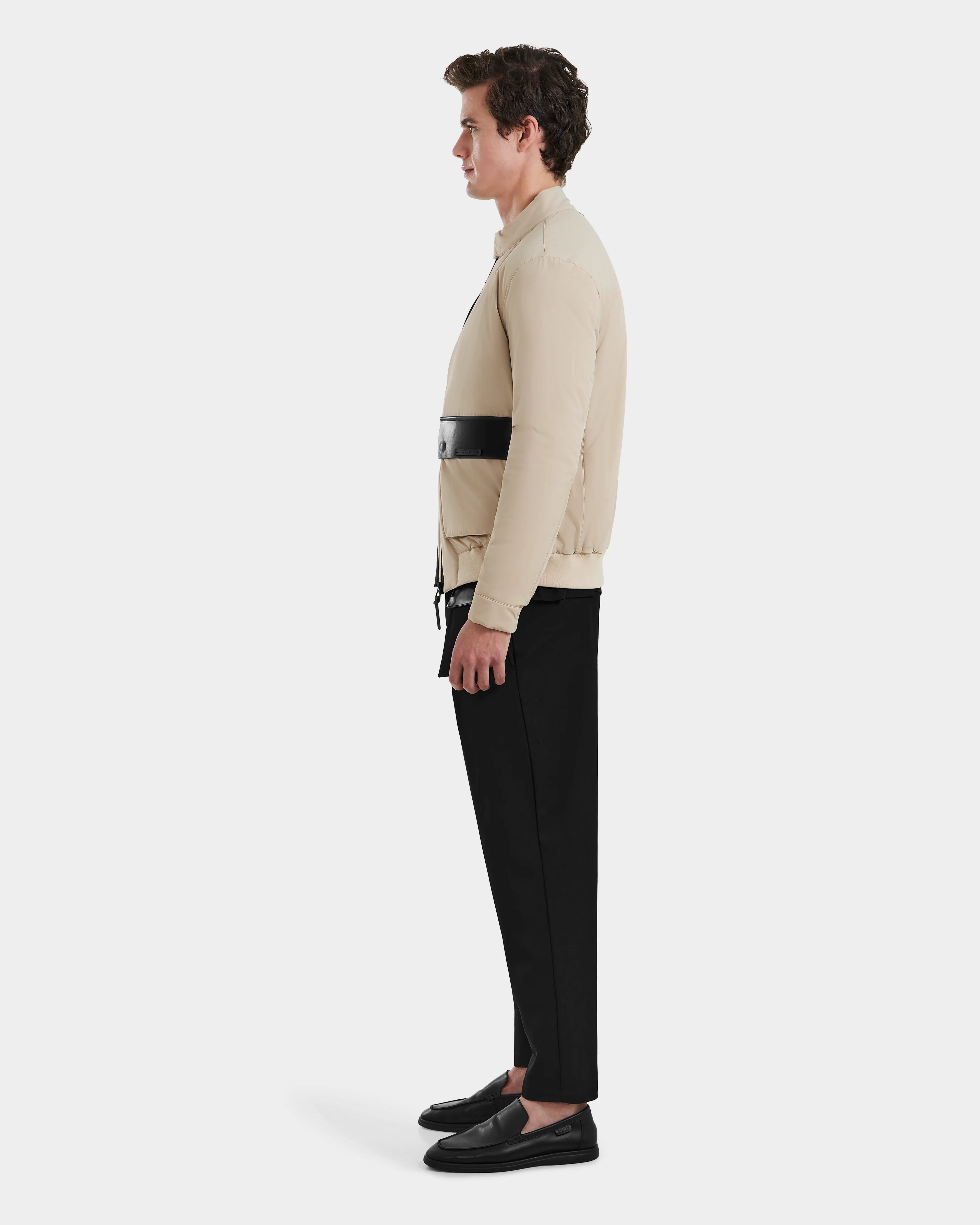 A man in profile wears the Rudsak PETER - 6125001 Almond bomber jacket—beige with a black horizontal stripe—paired with black pants and slip-on shoes against a plain white background.