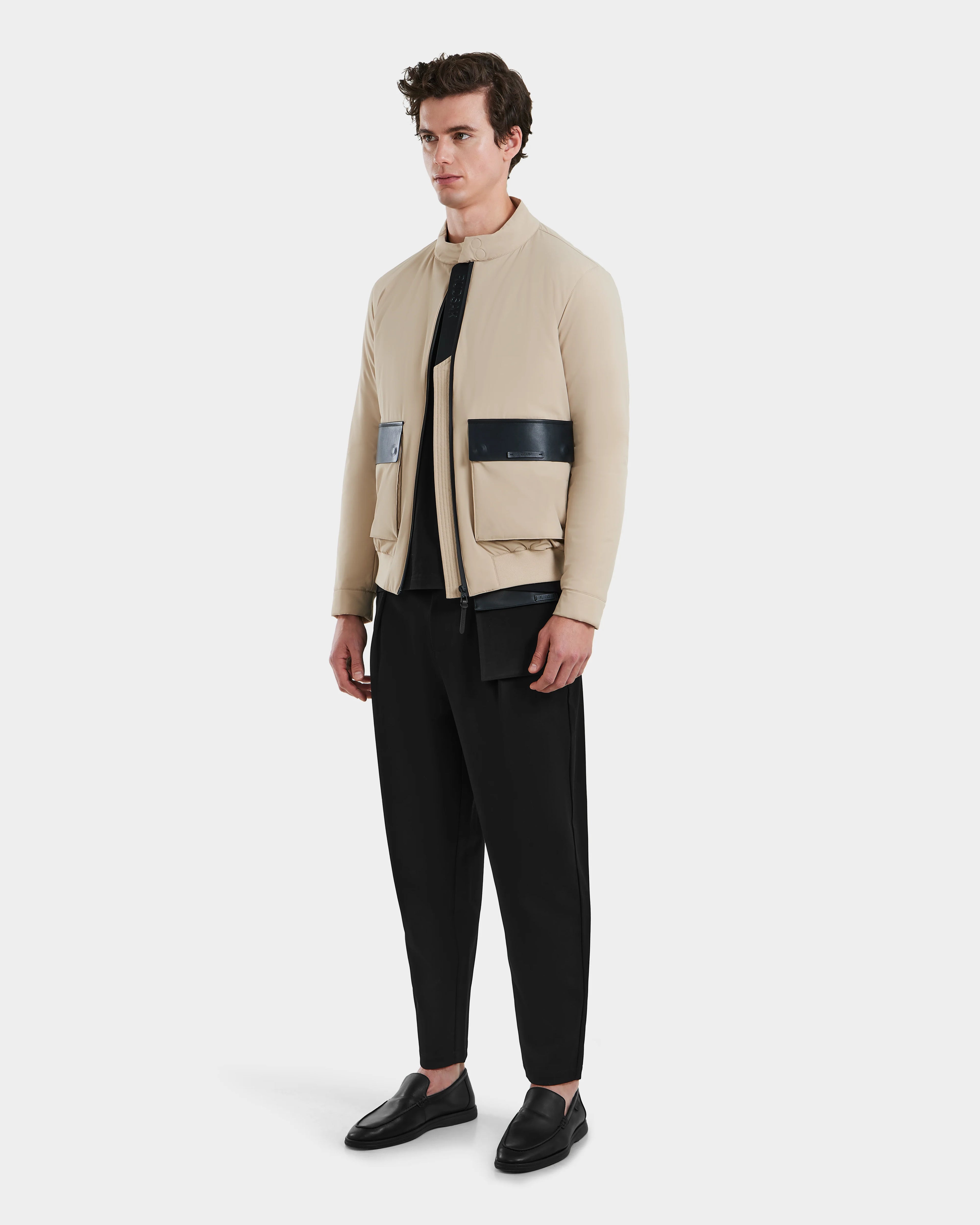 A man stands against a plain light background wearing the Rudsak PETER - 6125001 Almond beige lightweight bomber jacket with vegan leather accents, paired with a black shirt, loose black pants, and black loafers.