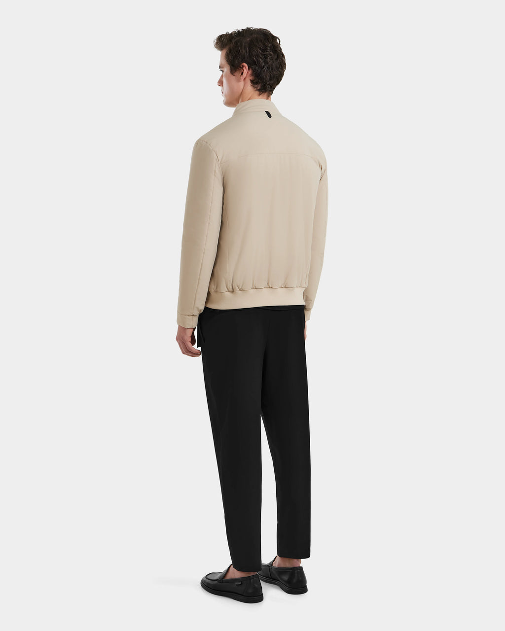 A man faces away wearing Rudsaks PETER - 6125001 Almond lightweight bomber with vegan leather accents, paired with black pants and shoes, against a plain white background.