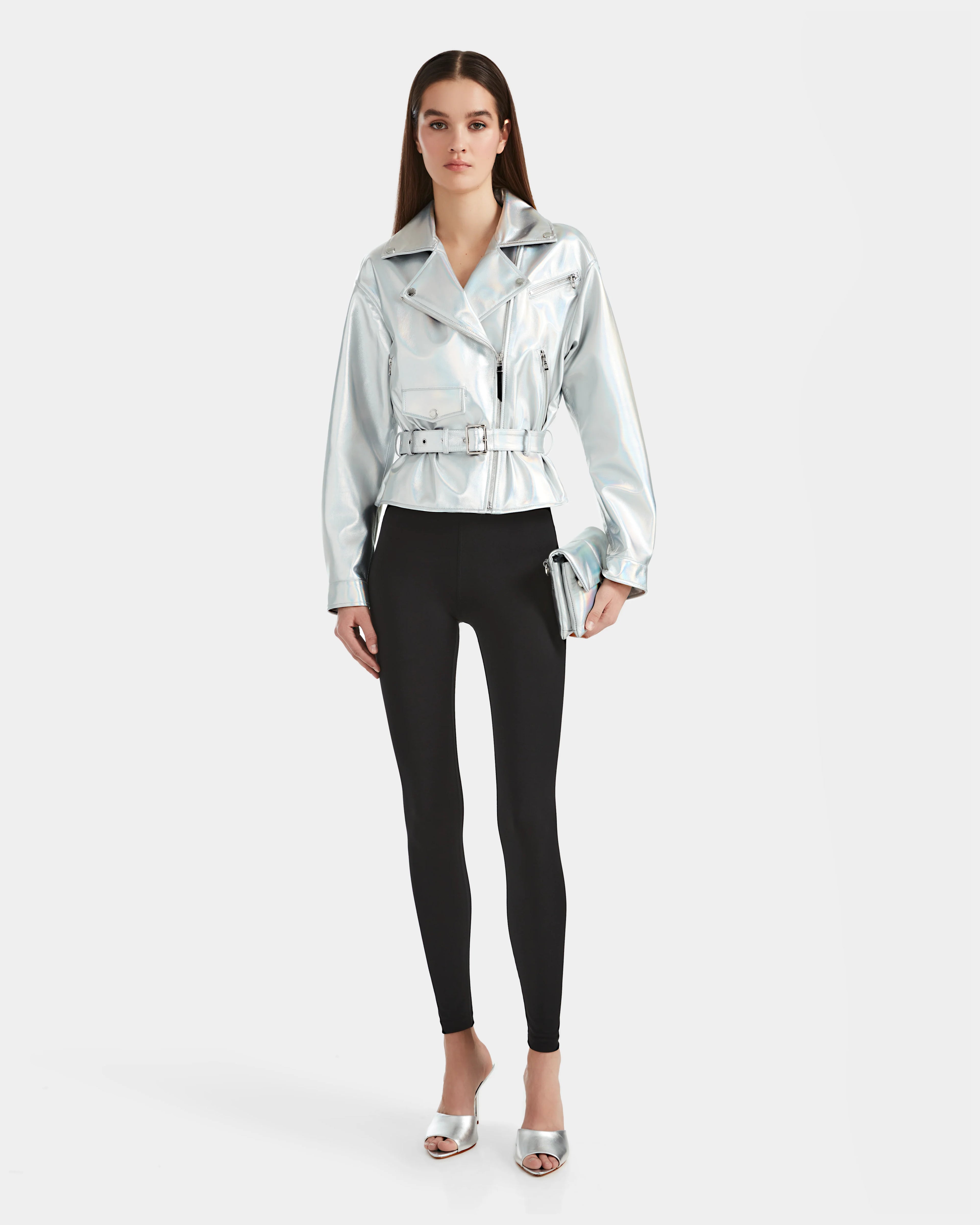 A woman with straight long hair and a neutral expression stands against a plain background wearing the Rudsak ARIELLE - 8124052 Iridescent Cotton White jacket, black leggings, silver open-toe heels, and holding a silver clutch.