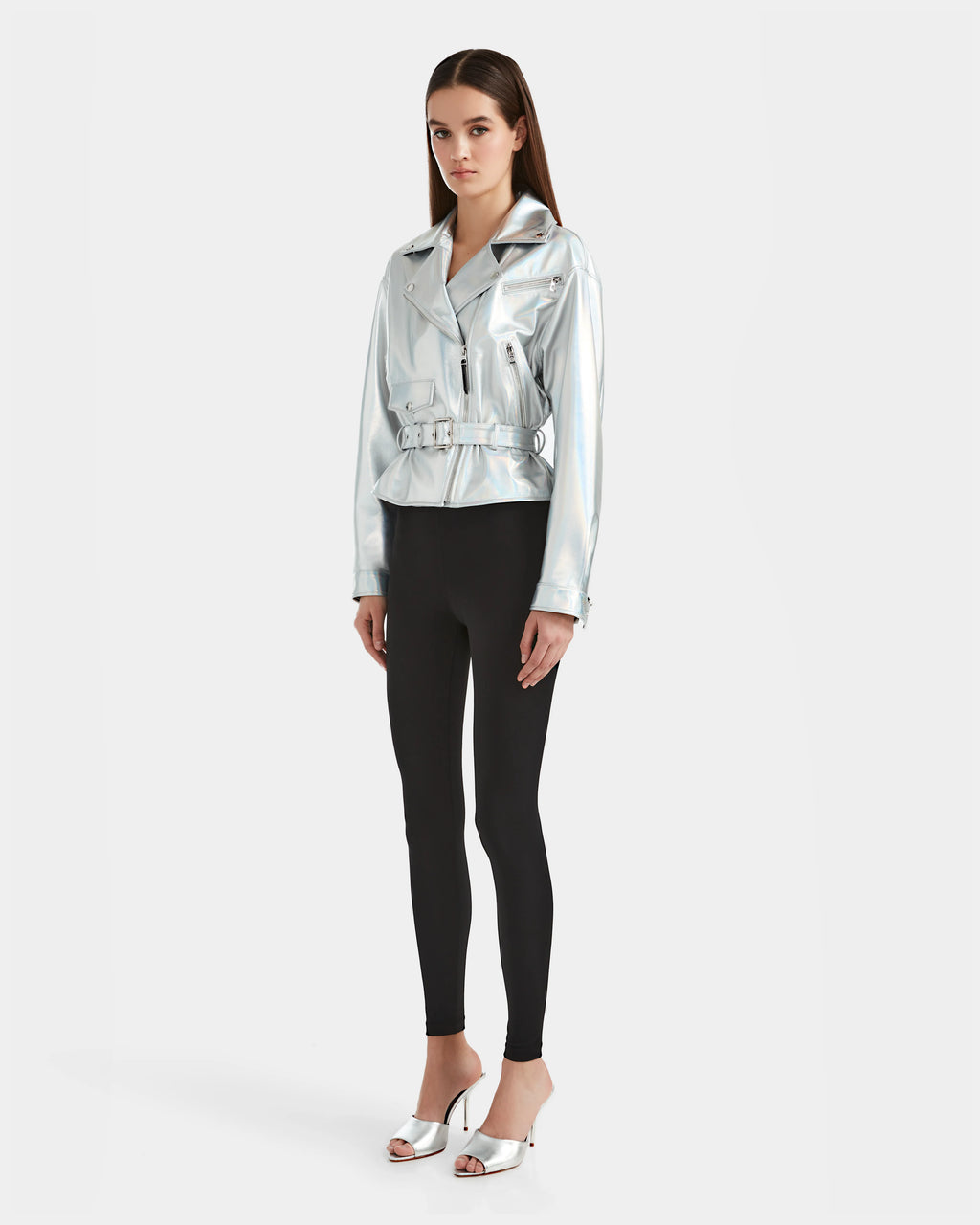 A woman with long straight hair and a neutral expression stands against a plain white background, wearing the Rudsak ARIELLE - 8124052 Iridescent Cotton White jacket with black leggings and silver open-toe heeled sandals.