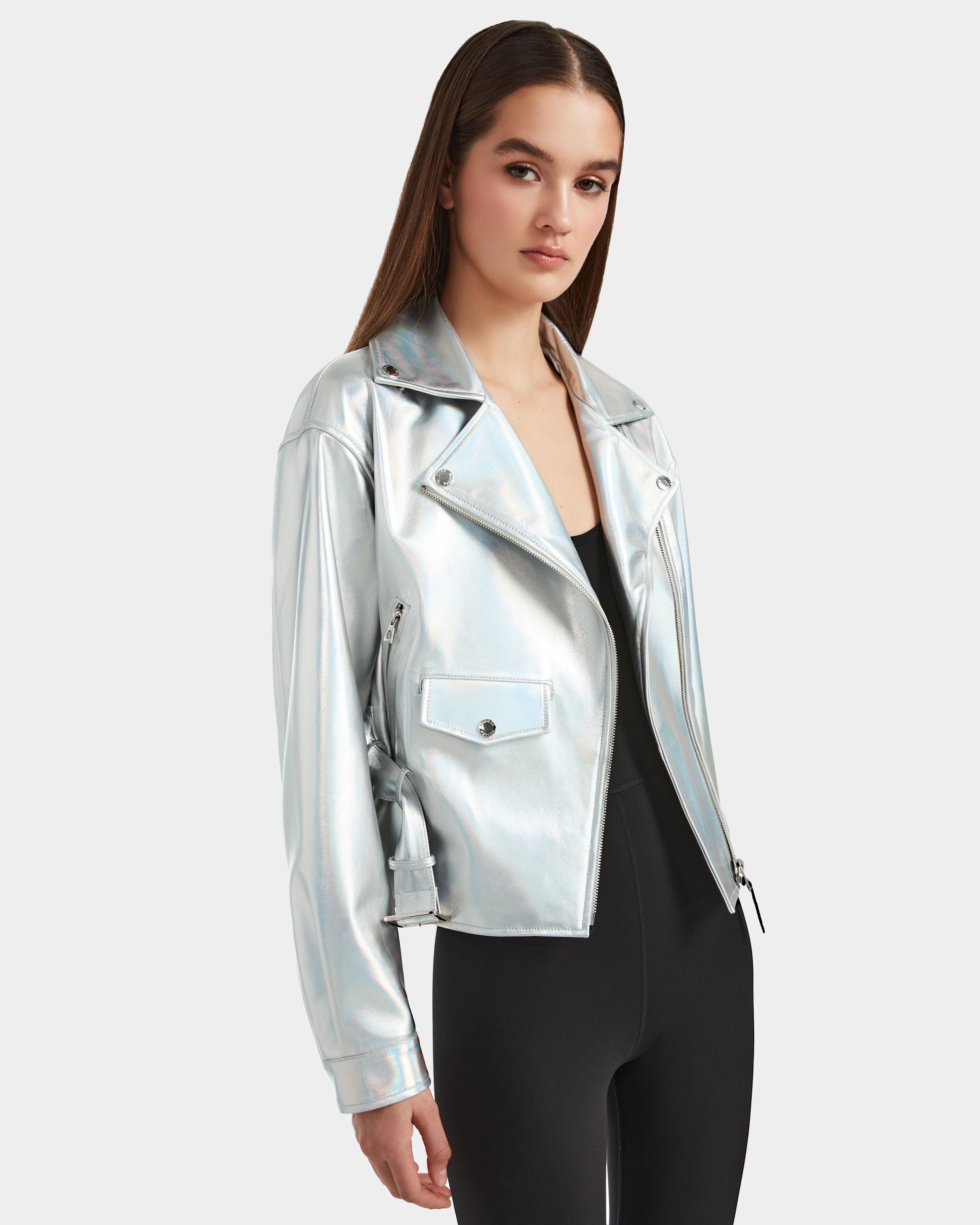 A woman with long brown hair wears the Rudsak ARIELLE - 8124052 Iridescent Cotton White biker jacket over a black outfit, standing against a plain light gray background.