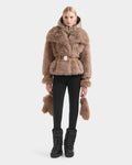 A woman with straight blonde hair and a neutral expression models the Rudsak CHLOE - 8125619 Camel faux fur jacket, featuring a matching belt and accents, paired with black leggings and boots against a plain white background.