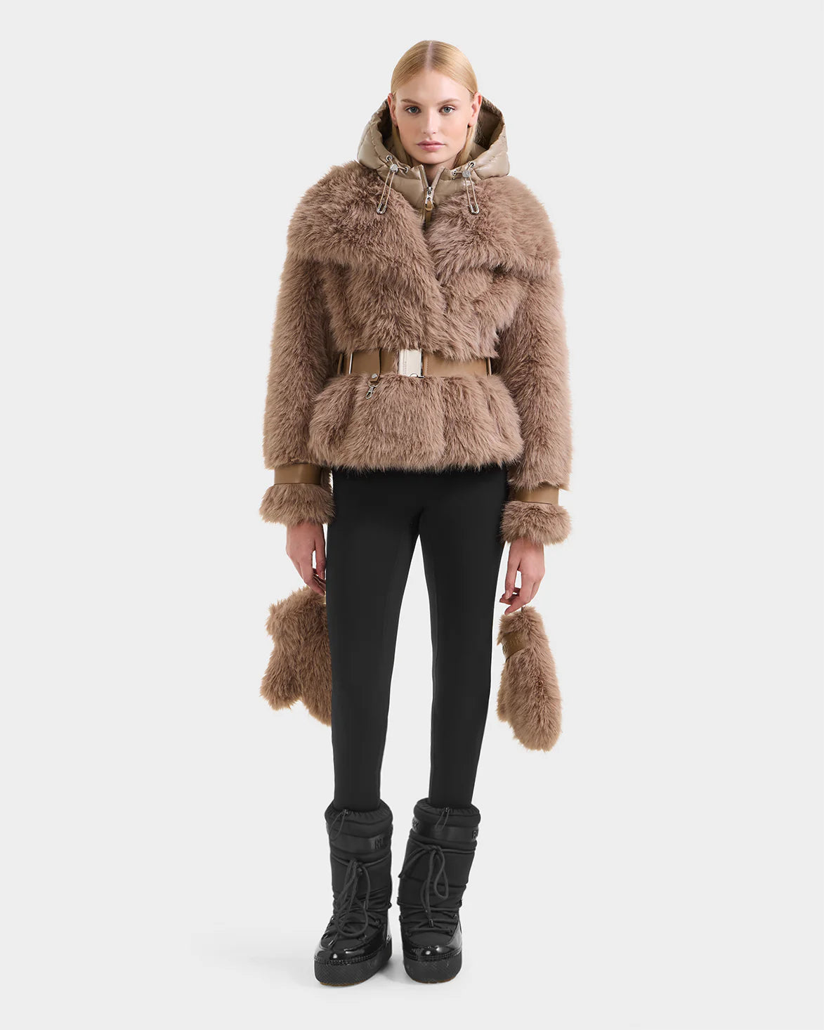 A woman with straight blonde hair and a neutral expression models the Rudsak CHLOE - 8125619 Camel faux fur jacket, featuring a matching belt and accents, paired with black leggings and boots against a plain white background.