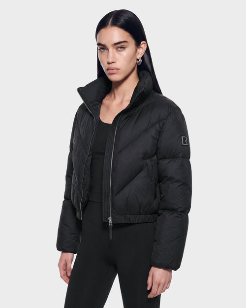 A woman with long dark hair stands against a plain light background, looking at the camera with a neutral expression. She is wearing the Rudsak ANSLIE - 8126986 Black puffer jacket and black pants.