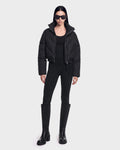 A person stands against a plain background wearing the Rudsak ANSLIE - 8126986 Black puffer jacket, paired with a black top, black leggings, knee-high black boots, and dark sunglasses.