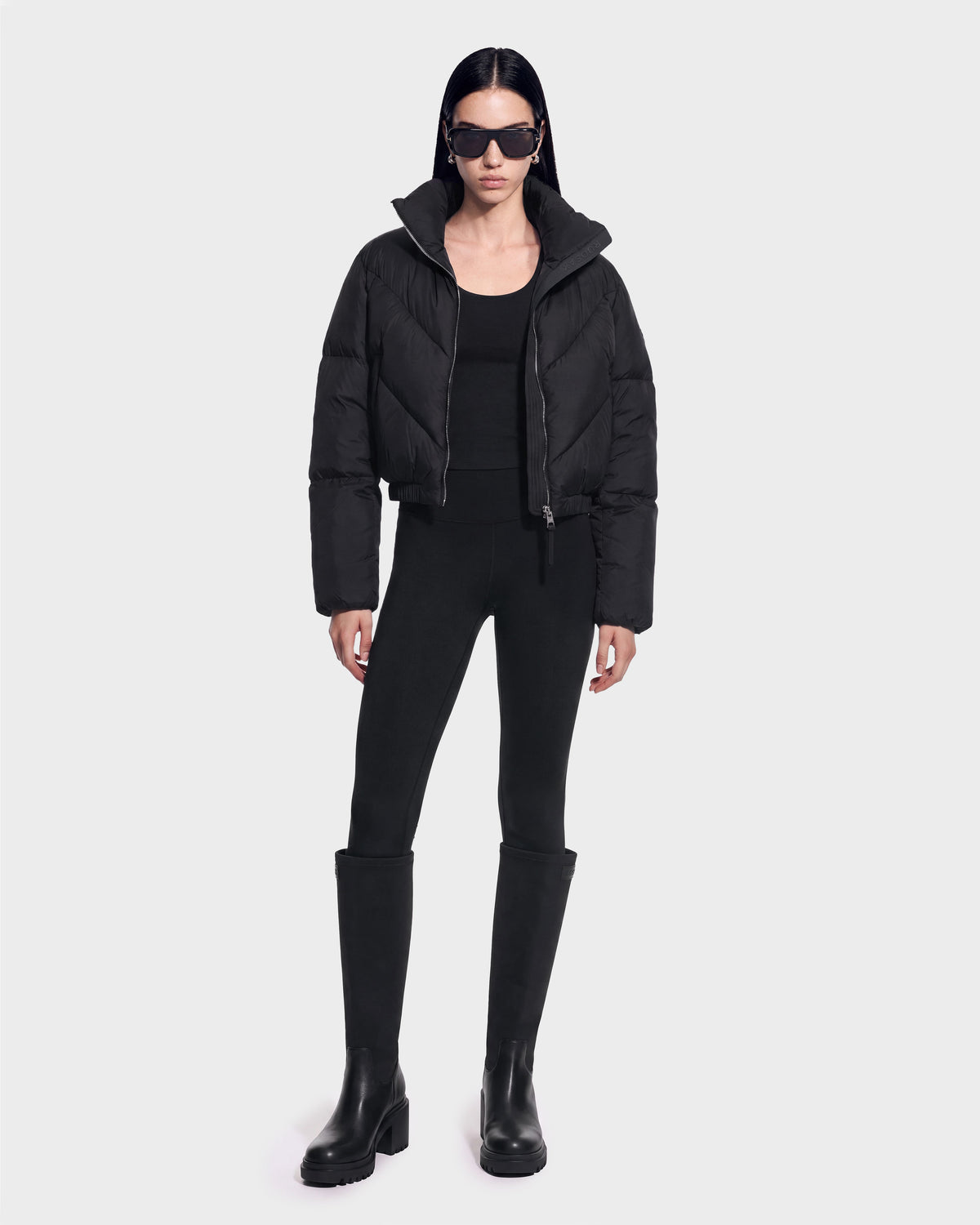 A person stands against a plain background wearing the Rudsak ANSLIE - 8126986 Black puffer jacket, paired with a black top, black leggings, knee-high black boots, and dark sunglasses.