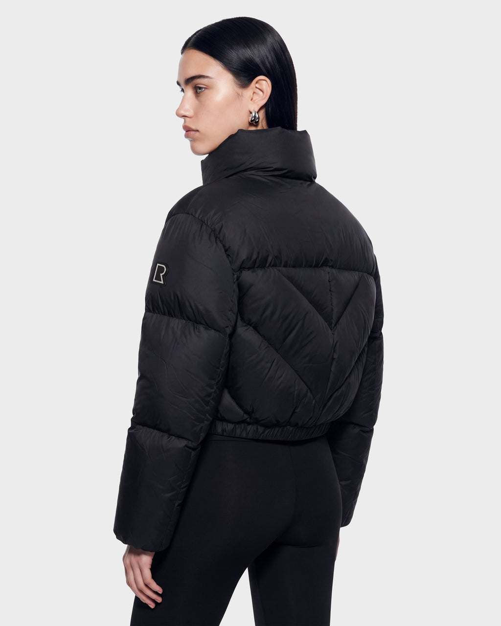 A woman with straight dark hair models the Rudsak ANSLIE - 8126986 Black puffer jacket, featuring a high collar and fitted black pants, as she stands against a plain light background, facing away while turning her head to the side.